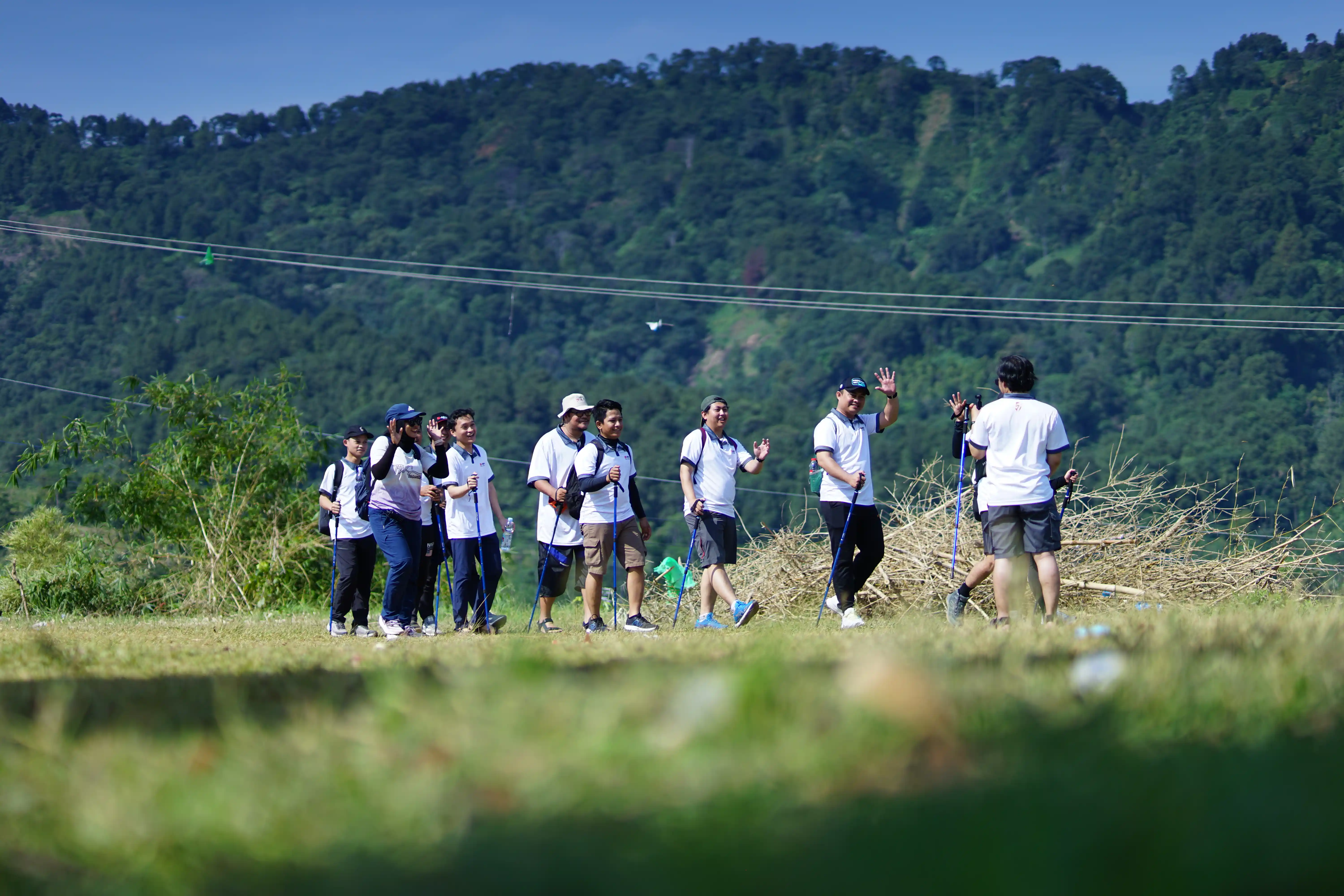 Rombongan peserta trekking berjalan menyusuri jalur setapak di antara semak belukar hijau.