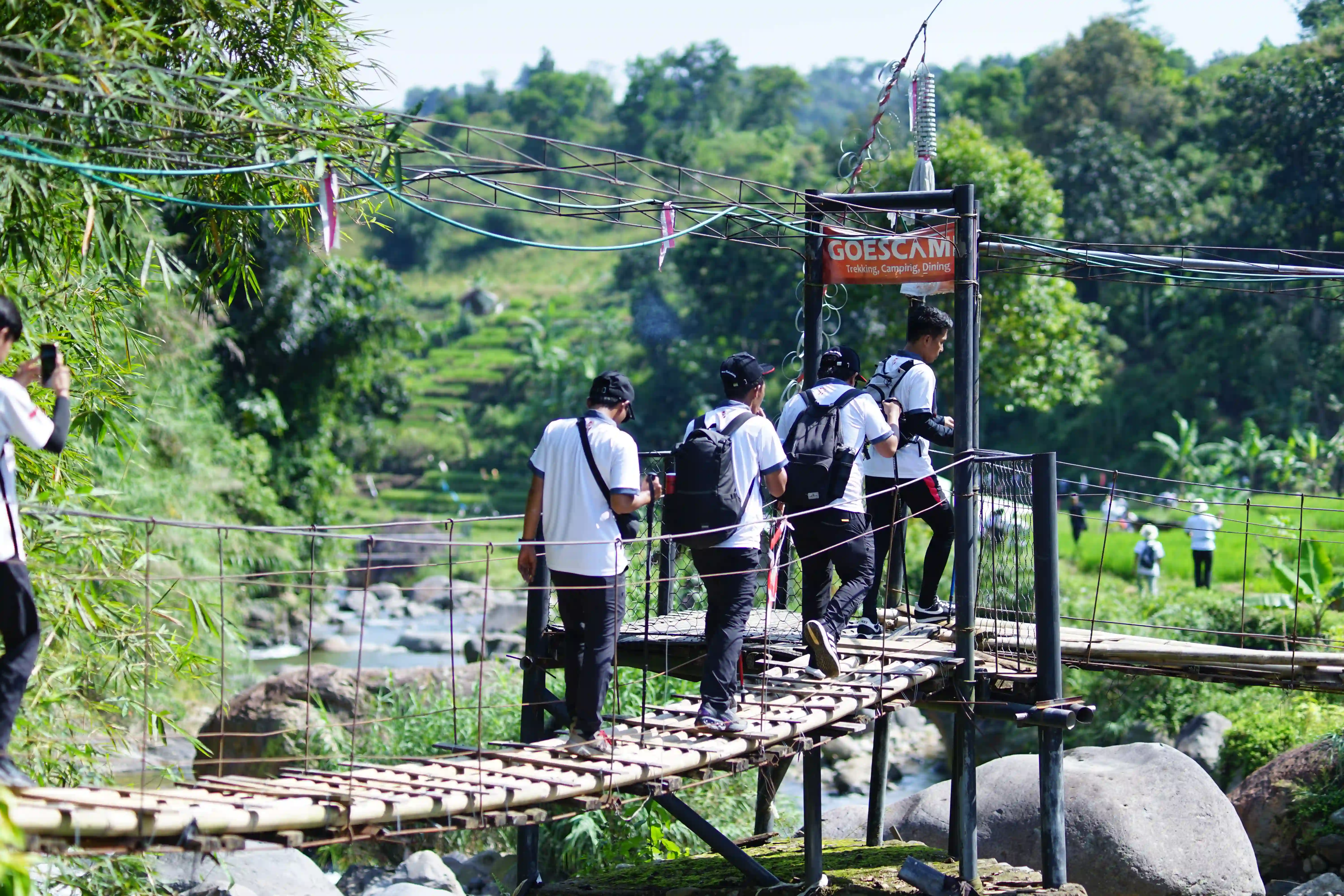 Beberapa peserta menyeberangi jembatan gantung bambu di atas sungai berbatu.