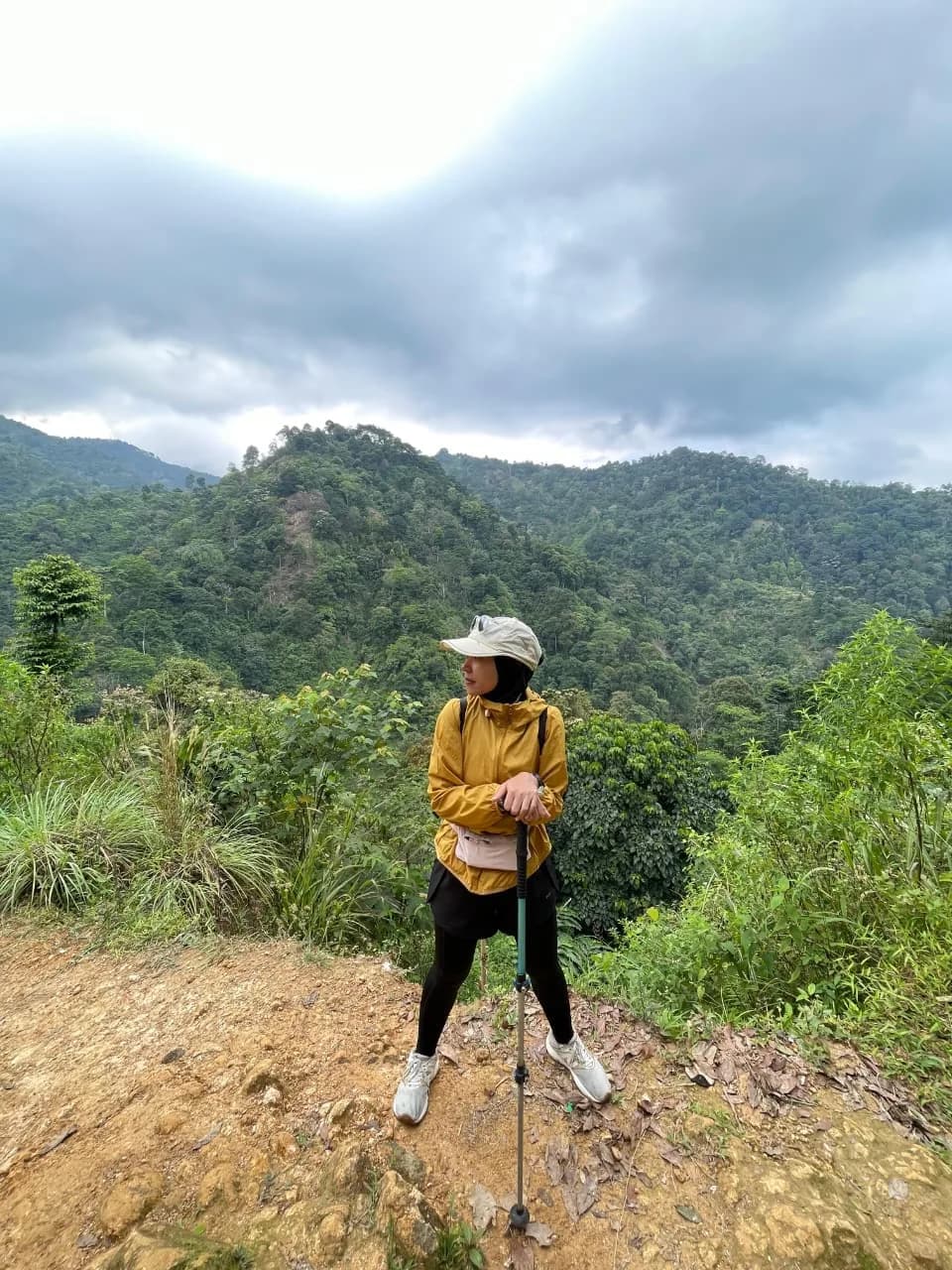 Seorang pendaki wanita menikmati pemandangan perbukitan hijau dari titik pandang.