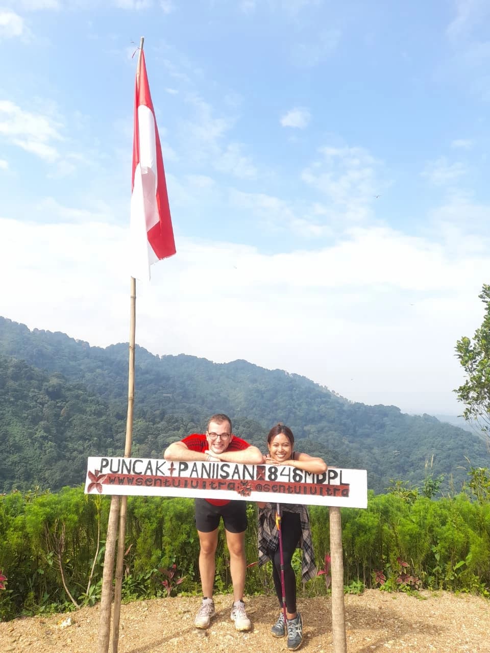 Pasangan pendaki berpose mesra di jalur trekking dengan latar belakang gunung yang hijau.